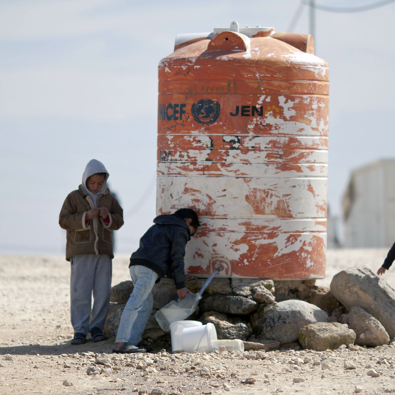 The image depicts children interacting near a large water tank. One child appears to be filling a container with water from a tap on the tank, while another child stands beside them. A third child is standing apart, looking at their phone or device. The water tank is orange and white, marked with a logo that indicates it is associated with a humanitarian organization. The surrounding area is dry and rocky, suggesting a challenging environment.