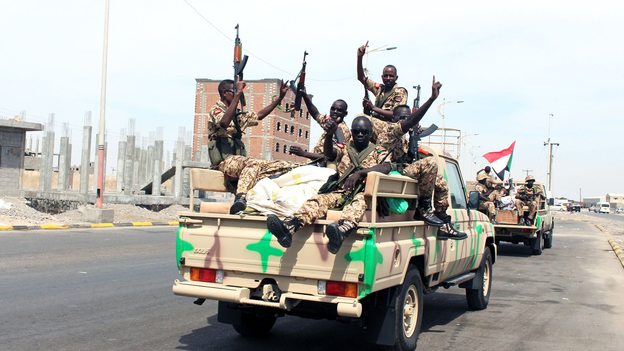 تظهر الصورة مجموعة من الجنود يجلسون على شاحنة عسكرية، حيث يحملون أسلحتهم ويرتدون زيًّا عسكريًا. يبدو أن الجنود في حالة احتفال أو انتصارات، حيث يُظهرون إشارات تحدٍ أو سعادة. الخلفية تحتوي على مبانٍ غير مكتملة، ما يشير إلى بيئة حضرية. الشاحنة تحمل ألوانًا عسكرية مموهة، مما يضيف إلى الطابع العسكري للصورة.