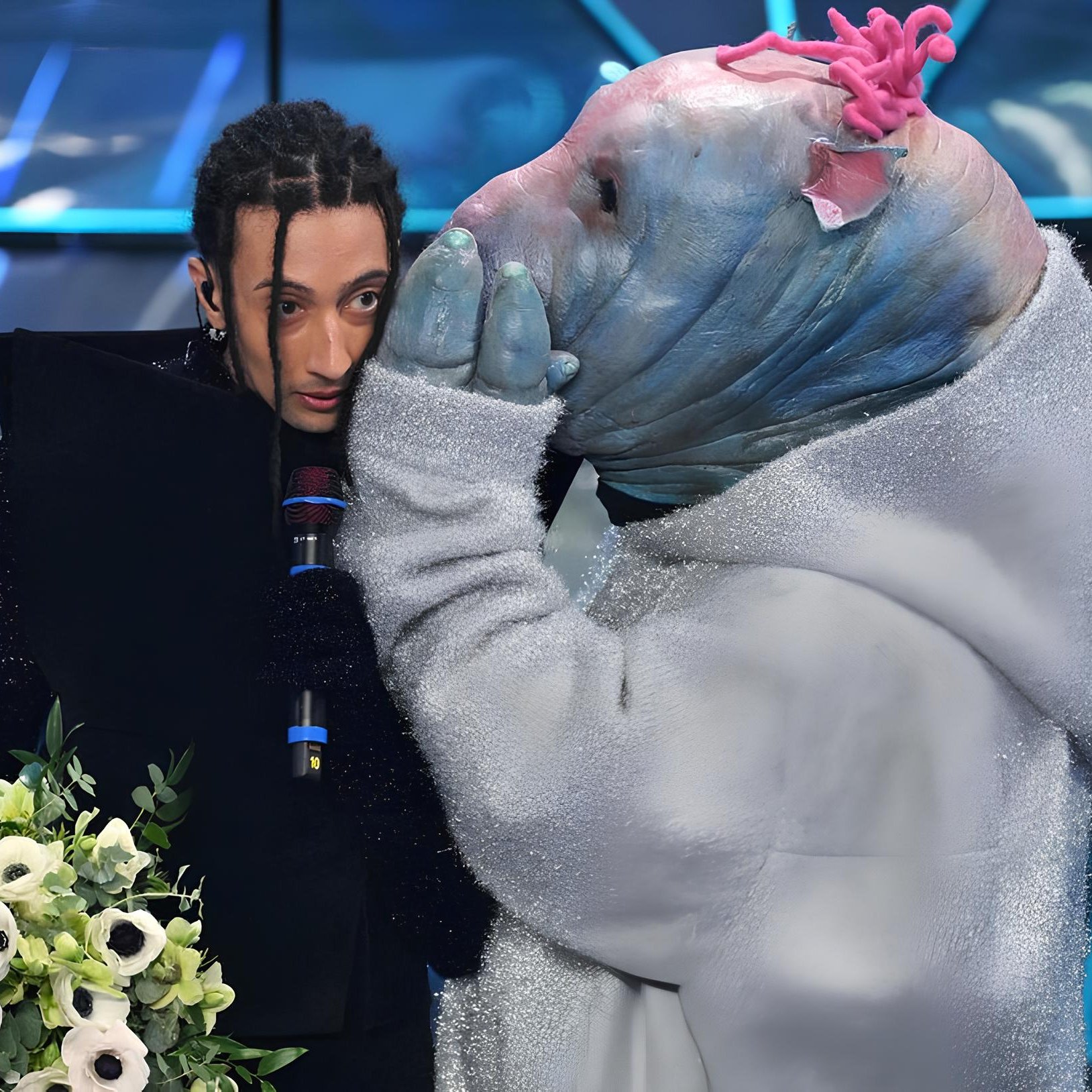 L'image montre une scène d'un spectacle où une personne au style vestimentaire unique, tenant un bouquet de fleurs, semble être en conversation avec une autre personne déguisée dans un costume original. Le costume ressemble à un animal aquatique, avec une tête d'apparence bleue et des détails colorés. L'ambiance semble dramatique ou émotive, avec un éclairage qui accentue les éléments de la scène.