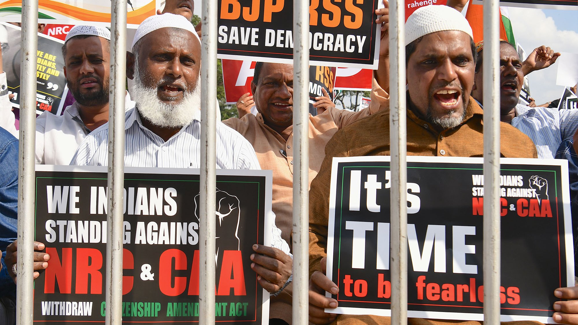 L'image montre un groupe de manifestants qui se tiennent derrière des barreaux. Ils tiennent des pancartes exprimant leur opposition à certaines lois et politiques en Inde, notamment le BJP, le NRC et le CAA. Les manifestants ont des expressions déterminées et sont clairement engagés dans leur cause, appelant à la démocratie et à la résistance. Les pancartes portent des messages en anglais et en d'autres langues, soulignant leur solidarité et leur demande de justice.