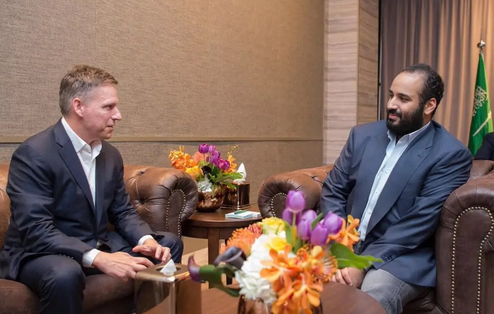 Deux hommes en costume assis dans un salon, entourés de fleurs colorées.