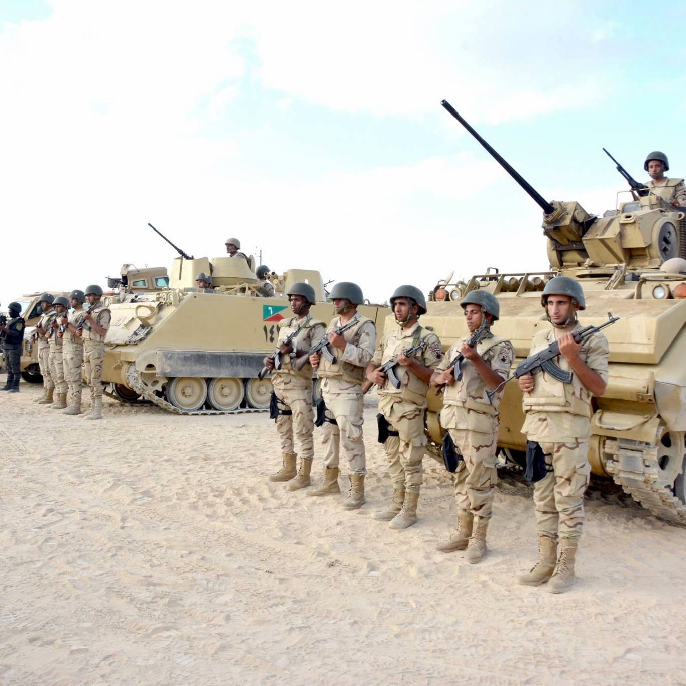 تظهر في الصورة مجموعة من الجنود العسكريين يقفون في صف منتظم، يرتدون زيًا عسكريًا ويحملون أسلحة. خلفهم توجد مدرعات عسكرية، وبعض الجنود يجلسون فوق هذه المدرعات. يبدو أن المشهد قد تم التقاطه في منطقة صحراوية أو رملية، مع سماء صافية في الخلفية. تعكس الصورة أجواءً عسكرية منظمّة وجادة.
