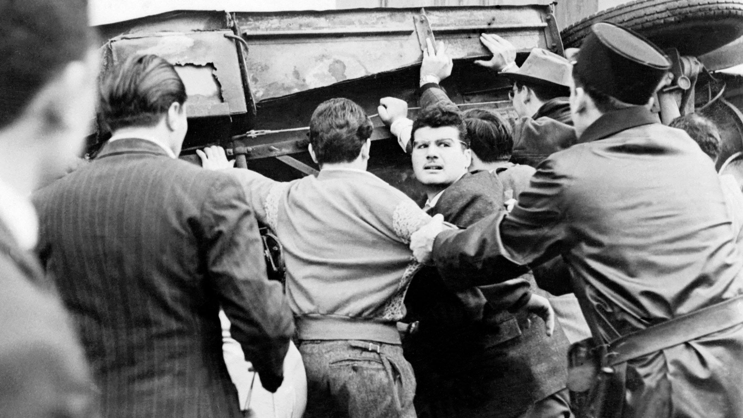 L'image montre une scène tumultueuse où un groupe de personnes semble être impliqué dans une altercation. Au centre, plusieurs hommes s'efforcent de soulever un véhicule renversé, tandis qu'un homme au visage concentré semble être en train de s'opposer à la situation ou d'interagir avec les autres. À droite, un policier en uniforme semble gérer la situation. L'atmosphère est tendue, et le contraste en noir et blanc accentue l'intensité de la scène.