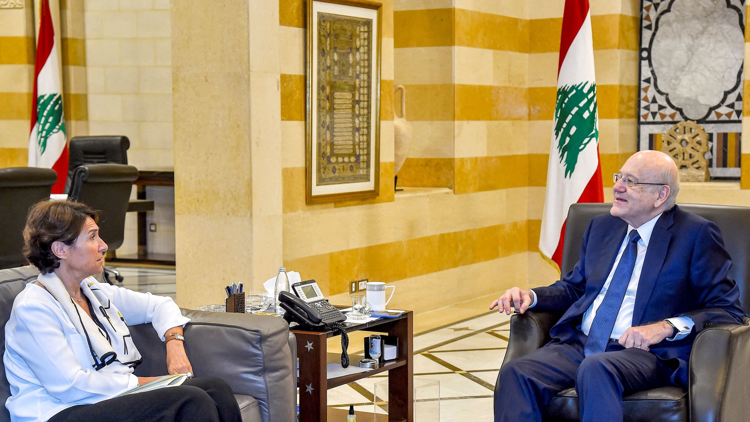 الصورة تظهر اجتماعًا داخل مكتب. يجلس رجل وامرأة على أريكتين. الرجل يبدو في منتصف العمر ويرتدي بدلة رسمية. المرأة ترتدي ملابس عادية، ويبدو أنها تشارك في الحوار. في الخلفية، تُرى ألوان العلم اللبناني، مما يدل على أن الاجتماع قد يكون ضمن إطار حكومي أو رسمي. الأثاث والتصميم الداخلي يعبّران عن الفخامة.