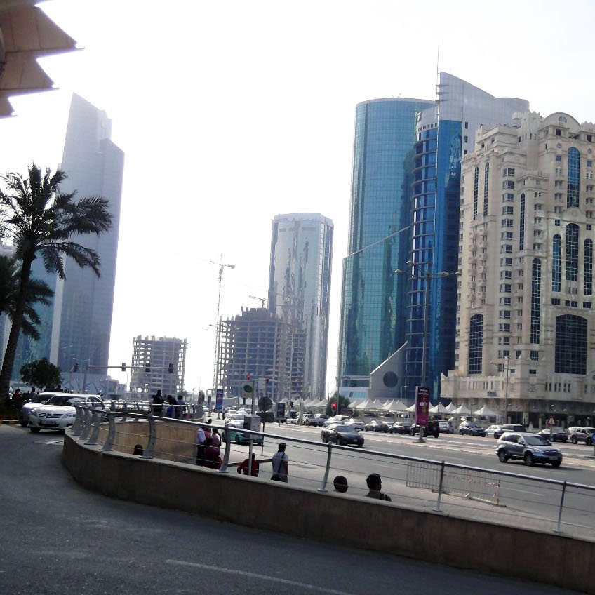 L'image montre une vue urbaine d'une ville moderne, avec des gratte-ciels en arrière-plan. On peut voir des bâtiments de différents styles architecturaux, certains sont très contemporains avec des façades en verre. Il y a des palmiers le long de la route, et on aperçoit des voitures circulant sur une avenue large. L'éclairage est clair, suggérant une journée ensoleillée. La scène donne une impression de dynamisme et de modernité.