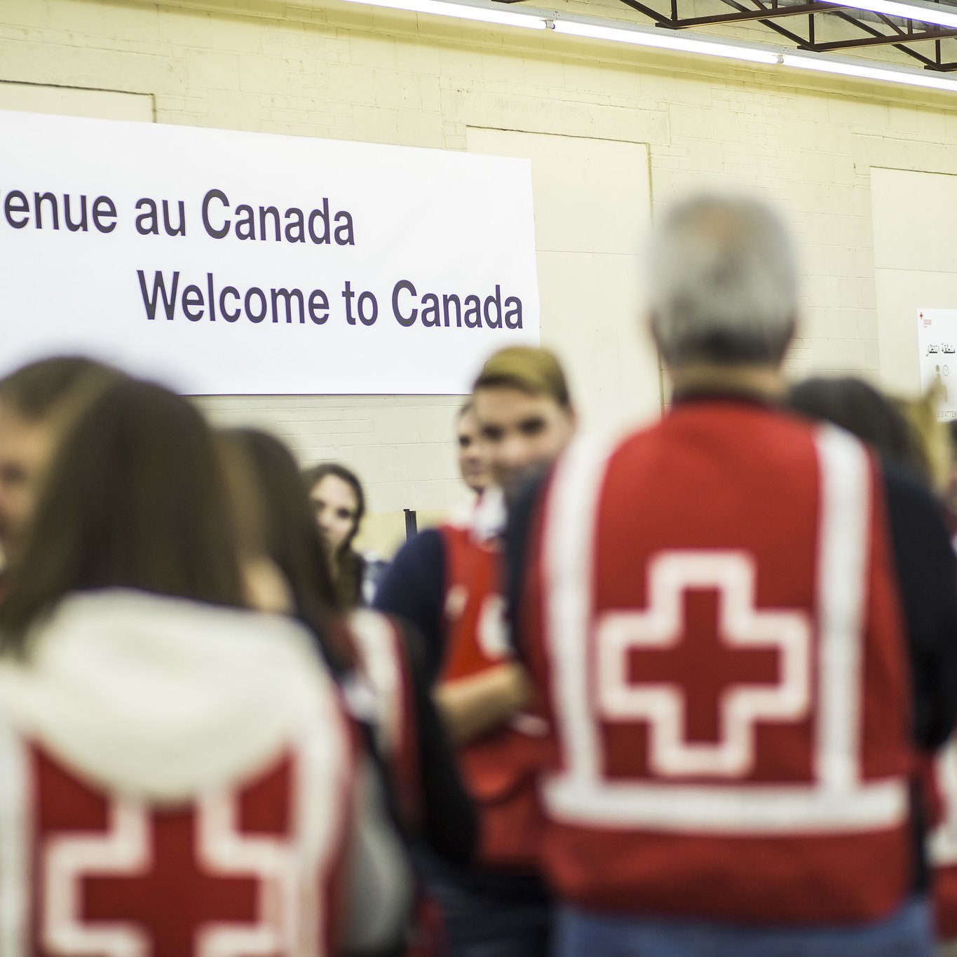 L'image montre un intérieur où un groupe de personnes se rassemble. Au fond, il y a une grande affiche indiquant "Bienvenue au Canada" en français et en anglais. Plusieurs personnes portent des vestes rouges avec une croix blanche, ce qui suggère qu'elles sont des secouristes ou des membres d'une organisation humanitaire. L'ambiance semble accueillante et axée sur l'accueil des nouveaux arrivants. Des drapeaux canadiens sont également visibles dans la scène.
