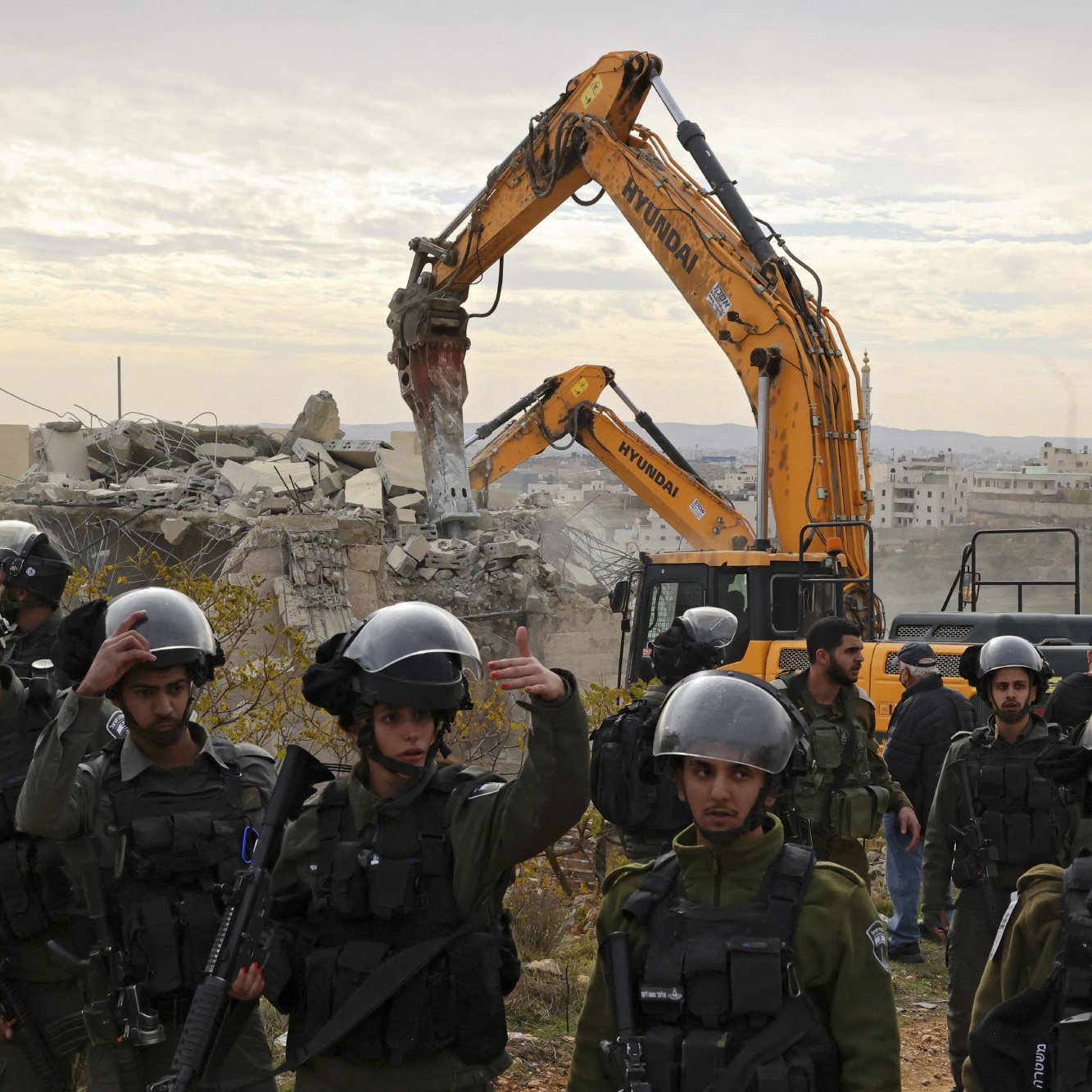 The image depicts a scene of destruction where a construction or demolition vehicle is actively tearing down a building, surrounded by a group of armed soldiers. The soldiers are wearing helmets and tactical gear, indicating a heightened security context. The background features a landscape with additional buildings, suggesting a possibly urban or conflict-affected area. The atmosphere appears tense, reflecting the serious nature of the situation being portrayed.