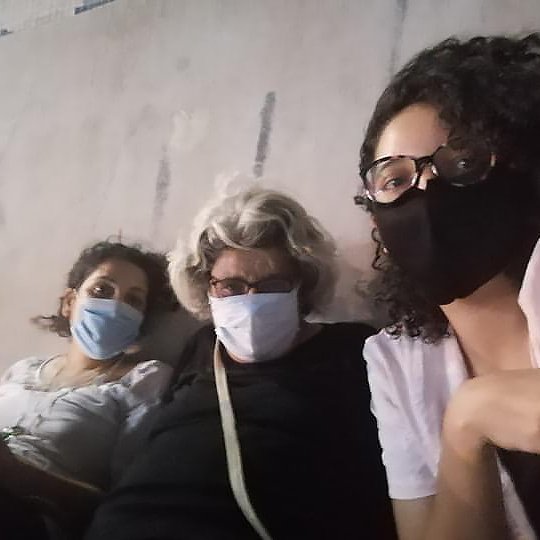 Sur l'image, on peut voir trois personnes assises côte à côte, chacune portant un masque. La première personne à gauche a les cheveux bouclés et porte un haut clair. La personne au milieu a des cheveux gris et porte des lunettes ainsi qu'un haut noir. La dernière personne à droite a des cheveux bouclés et porte également un haut clair. Elles semblent se détendre ensemble, peut-être dans un environnement public ou en extérieur. L'ambiance est informelle et conviviale.