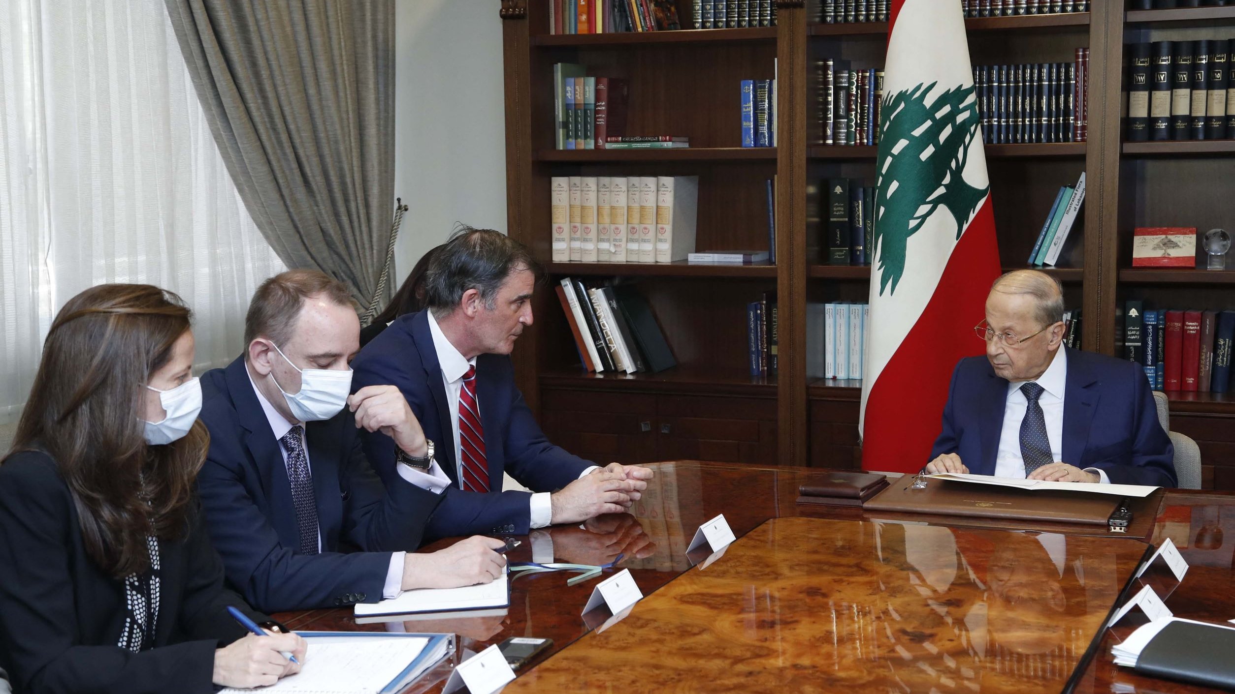 Nell'immagine, si può vedere un incontro ufficiale in una sala ben arredata. Ci sono quattro persone sedute attorno a un grande tavolo in legno. A destra, un uomo anziano, presumibilmente un leader o un ufficiale governativo, è rivolto verso gli altri partecipanti. Gli altri tre, seduti a sinistra, indossano mascherine e stanno prendendo appunti su quaderni. Dietro di loro, si intravede una libreria piena di libri e una bandiera del Libano. L'atmosfera sembra seria e professionale, tipica di una discussione di Stato.