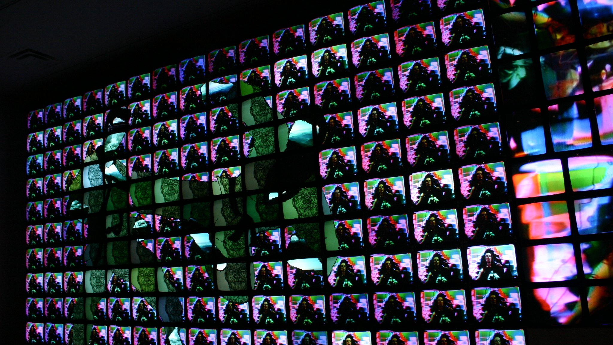 L'image présente une installation artistique composée de nombreux écrans alignés de manière à former un tableau visuel. Chaque écran affiche des images colorées, vibrantes et abstraites, créant un effet d'ensemble dynamique. Les couleurs se mélangent et se chevauchent, ajoutant une sensation de mouvement. La composition semble jouer avec la lumière et les contrastes, attirant l'œil vers des motifs diversifiés. L'ensemble évoque une immersion dans une expérience numérique moderne et artistique.