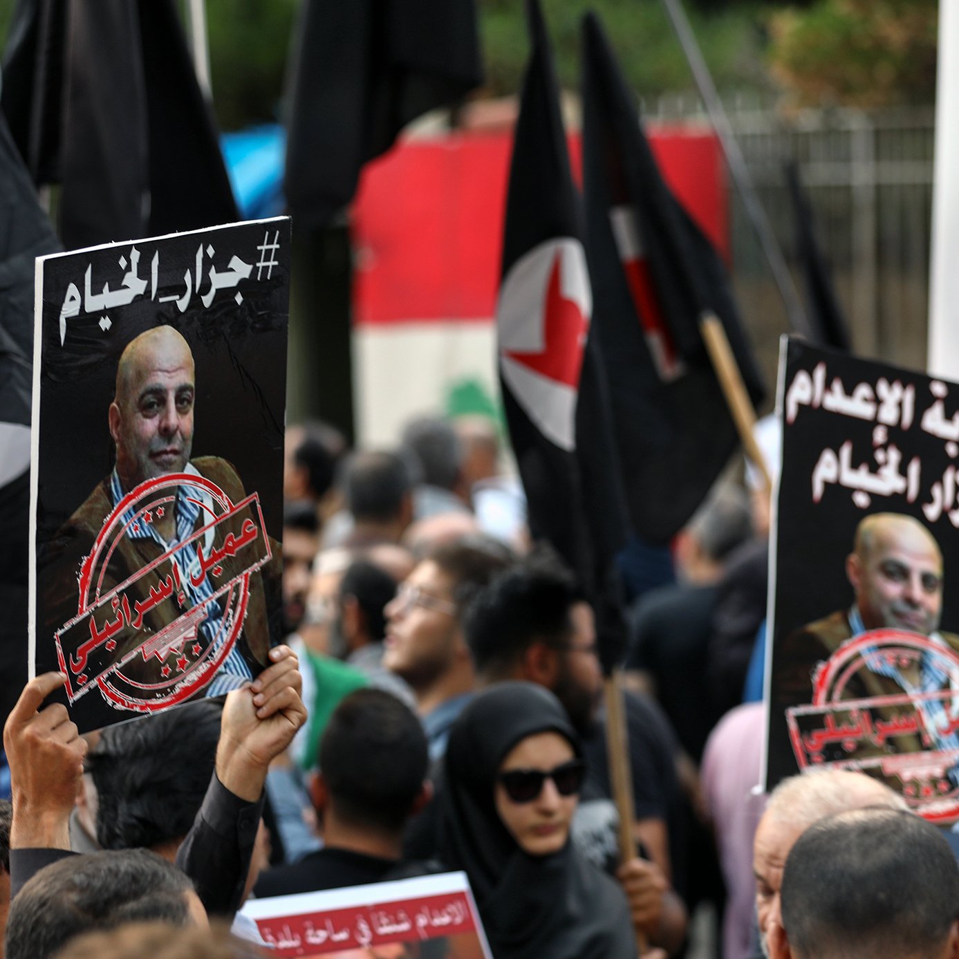 L'image montre une manifestation avec des personnes tenant des pancartes et des drapeaux. Les pancartes contiennent des portraits de personnes et des textes en arabe, évoquant des messages politiques ou sociaux. La foule semble engagée dans une cause, exprimant des émotions fortes. On peut voir une diversité de participants, certains portant des vêtements traditionnels et d'autres plus modernes, ainsi qu'un fond urbain suggérant un rassemblement public.