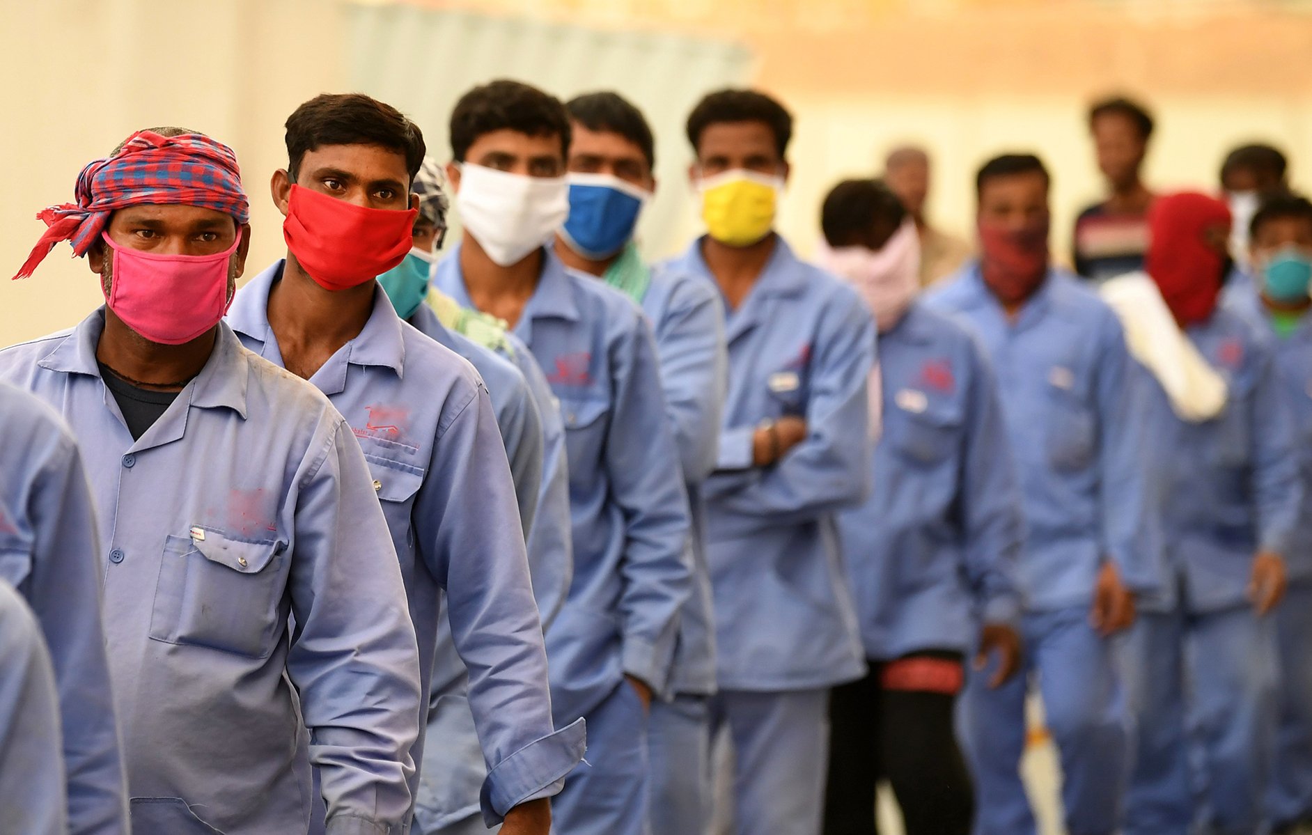 L'image montre un groupe de travailleurs masculins alignés en rang. Ils portent tous des vêtements de travail de couleur bleue et des masques de protection de différentes couleurs, notamment rouge et jaune. Les hommes semblent se préparer à entrer quelque part, et l'ambiance générale indique un contexte de travail, possiblement dans une situation où des mesures sanitaires sont en place. L'arrière-plan est flou, mettant l'accent sur les travailleurs.