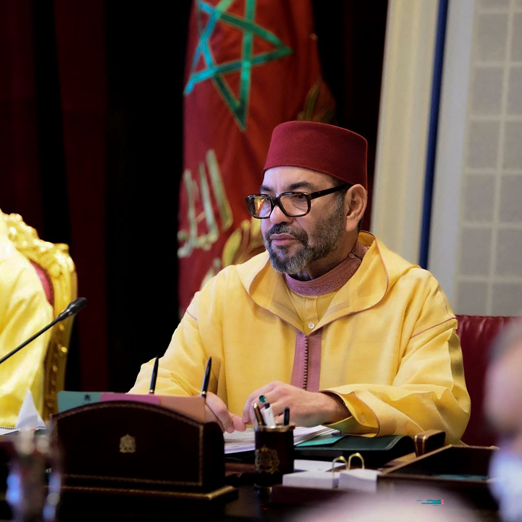 L'immagine mostra un incontro ufficiale in corso. A sinistra, un giovane indossa un abito giallo e una mascherina, mentre a destra c'è un uomo con un berretto rosso e un abbigliamento tradizionale marocchino di colore giallo. L'uomo a destra sembra concentrato, forse mentre prende appunti o partecipa a una discussione. Sullo sfondo si intravedono elementi decorativi di un ambiente formale.