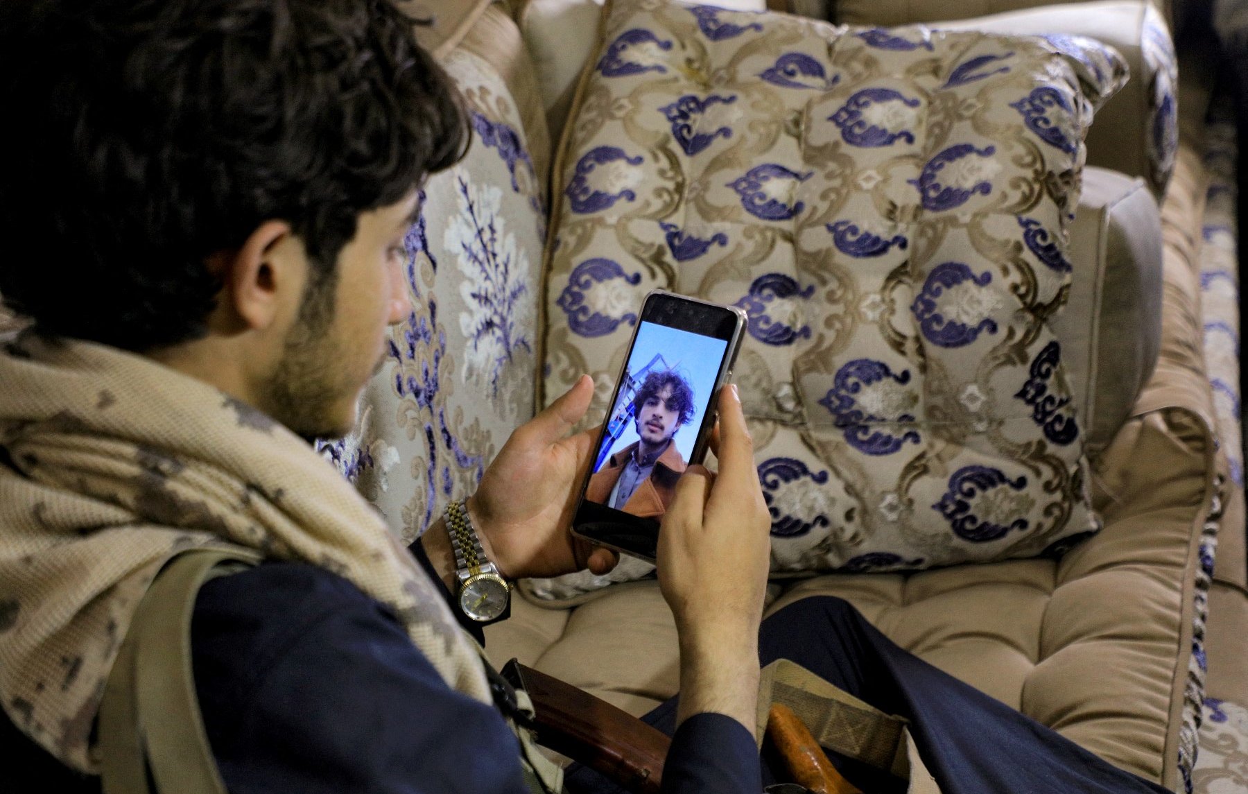 L'image montre un jeune homme assis sur un canapé décoré, regardant son téléphone portable. Il porte une écharpe et semble contemplatif. À côté de lui, on peut apercevoir une boîte et un objet, peut-être une arme ou un accessoire. Le décor est chaleureux et traditionnel, avec des coussins ornés de motifs élégants.