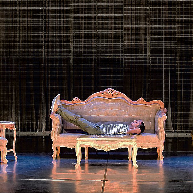 L'image montre une scène de théâtre avec un décor composé de plusieurs meubles anciens. Au centre, un sofa sur lequel un homme est allongé, semble détendu. À droite, une femme est assise sur une chaise, elle semble engagée dans une conversation ou une réflexion. Le fond est constitué d'un rideau sombre, ce qui met en valeur les meubles éclairés. L'ambiance semble à la fois intime et dramatique, suggérant une interaction entre les deux personnages.