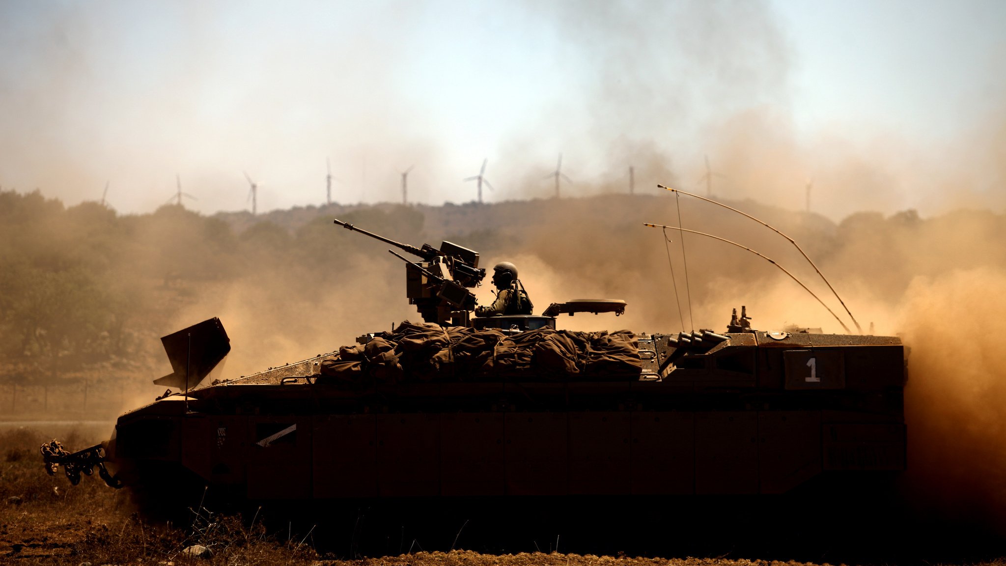 L'image montre un véhicule militaire, probablement un blindé, en action dans un paysage poussiéreux. Un soldat est installé sur le toit du véhicule, armé et prêt. En arrière-plan, on peut apercevoir des éoliennes qui se dressent contre un ciel clair. La scène évoque une ambiance de tension et de préparation militaire, tandis que la poussière soulevée par le mouvement ajoute une dynamique à l'image.