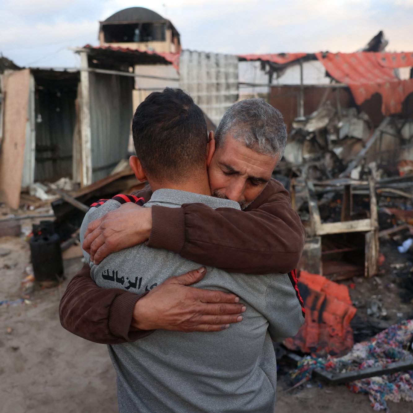 L'immagine mostra due uomini che si abbracciano in un contesto distrutto. Sullo sfondo, si possono vedere i resti di edifici danneggiati, con detriti e rovine sparse. La scena trasmette un forte senso di emozione e solidarietà tra le due persone, che si sostengono in un momento difficile. La luce naturale del cielo aggiunge un'atmosfera malinconica alla situazione.