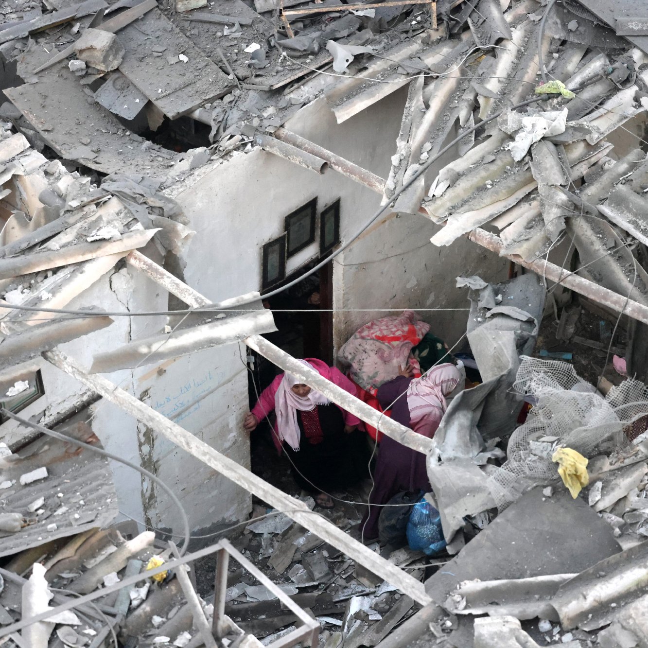 L'image montre des ruines d'un bâtiment, avec des débris de toit et des murs effondrés. Deux personnes sont visibles à l'intérieur des décombres, semblant chercher ou s'entraider dans cet environnement chaotique.