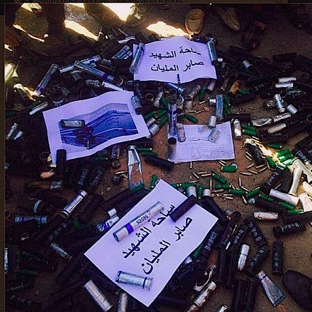 L'image montre un grand nombre de cartouches de munitions et de projectiles disposés au sol. Certaines de ces munitions sont accompagnées de panneaux ou de feuilles de papier avec des inscriptions en arabe. On peut voir des personnes autour de cette disposition, mais leurs visages ne sont pas visibles. L'ensemble de la scène semble évoquer une situation liée à la confiscation ou à la destruction d'armements.