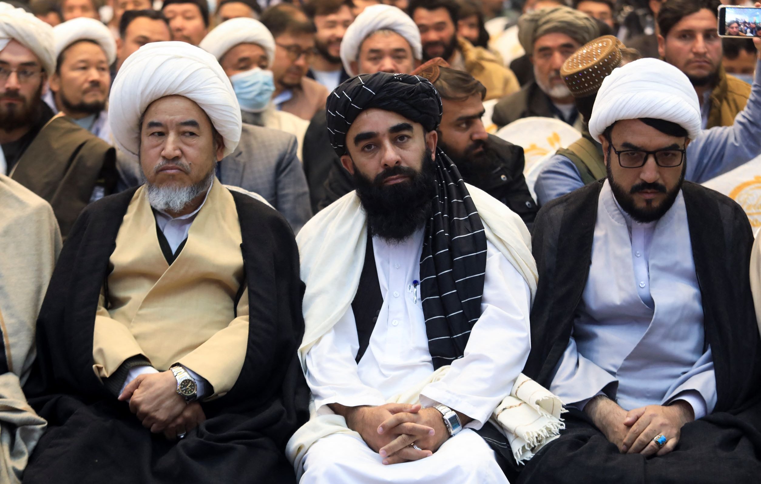 L'immagine mostra un gruppo di uomini seduti in un contesto di evento o incontro. I partecipanti indossano abiti tradizionali, tra cui chador e turbanti. I volti esprimono varie emozioni, alcuni sembrano seri e pensierosi. Sullo sfondo si possono vedere altre persone, suggerendo che si tratta di un'assemblea o di una manifestazione. L'atmosfera sembra essere formale e rispettosa.