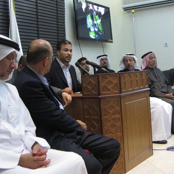 L'image montre un groupe de personnes rassemblées dans une salle. Au centre, un homme est en train de parler devant un microphone, tandis que les autres l'écoutent attentivement. Certains d'entre eux portent des vêtements traditionnels, comme des kandouras et des ghutras, indiquant peut-être un contexte culturel spécifique. À l'arrière-plan, un écran affiche probablement une présentation ou une vidéo. L'atmosphère semble sérieuse et concentrée, suggérant que la réunion pourrait porter sur un sujet important.