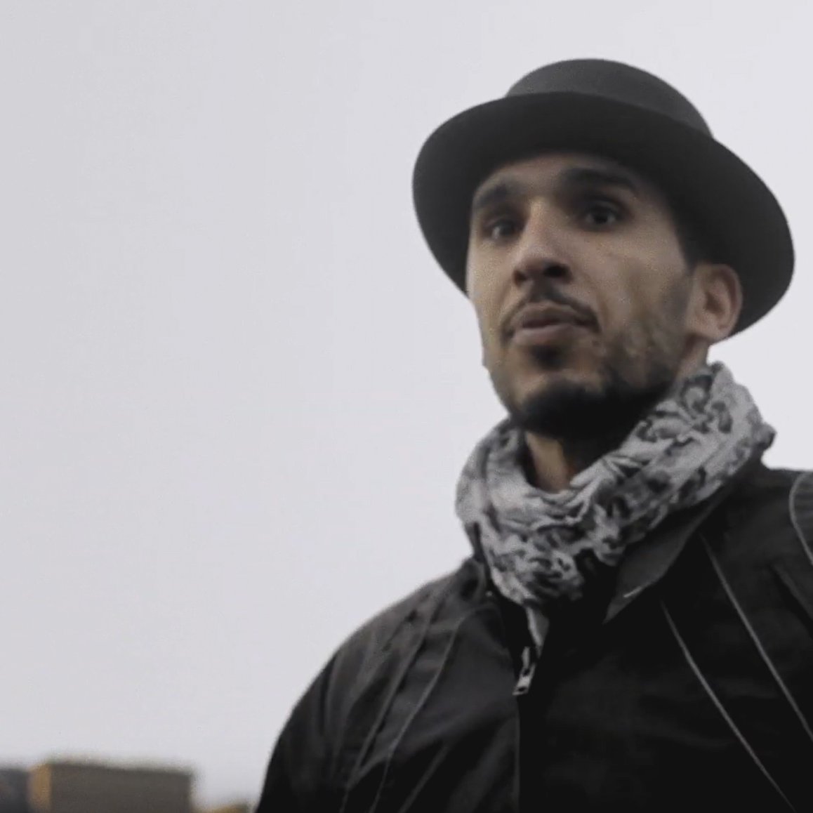 L'image montre un homme portant un chapeau noir et une écharpe. Il a une expression sérieuse et regarde droit devant lui. Le fond semble flou, suggérant une ambiance extérieure peut-être en milieu urbain ou dans un parc. Le temps semble nuageux, ce qui ajoute une atmosphère un peu sombre à la scène.