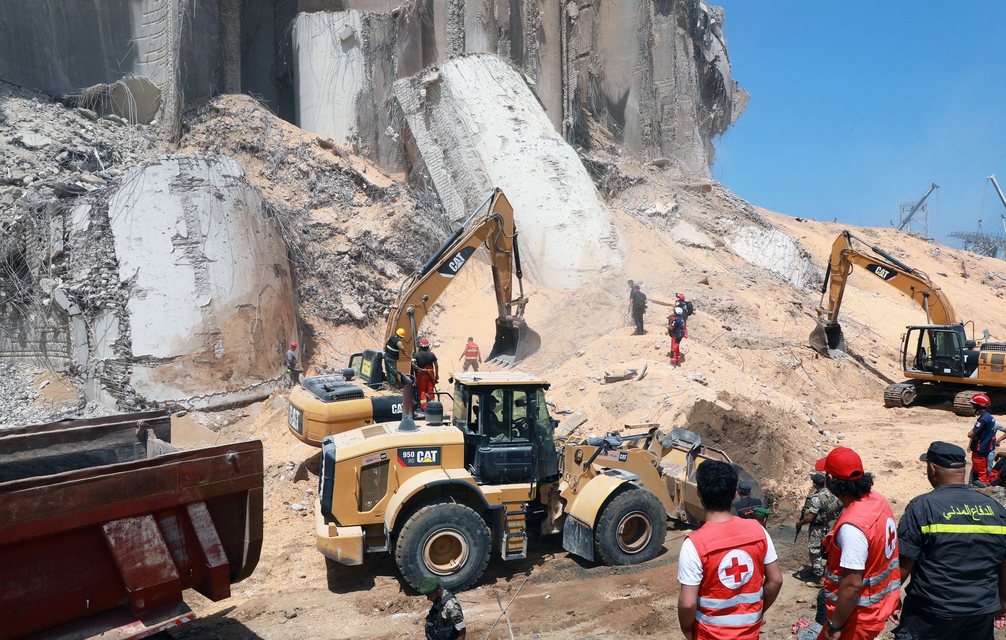 L'image montre un site de construction ou de déblaiement où des engins de chantier, tels que des pelles mécaniques et des bulldozers, sont en action. Au premier plan, des travailleurs en uniforme, y compris des membres de la Croix-Rouge, supervisent les opérations. En arrière-plan, on peut voir des débris et des structures effondrées, suggérant une situation d'urgence ou de nettoyage après un incident. Le ciel est dégagé et ensoleillé, ajoutant un contraste avec l'environnement de travail.