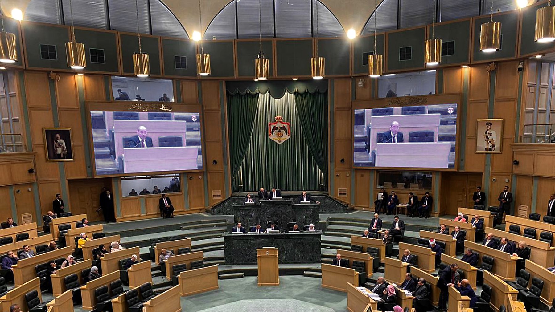The image depicts an interior view of a legislative assembly or parliament. The chamber is spacious, featuring a semi-circular arrangement of desks for members. At the front, there is a podium where a speaker or official is addressing the assembly. Large screens on either side display information or the speaker to the attendees. The walls are adorned with drapes and emblems, contributing to a formal atmosphere. Members of the assembly are seated, engaged in discussion or listening attentively. The overall setting is one of a governmental deliberation space, likely during a session or meeting.