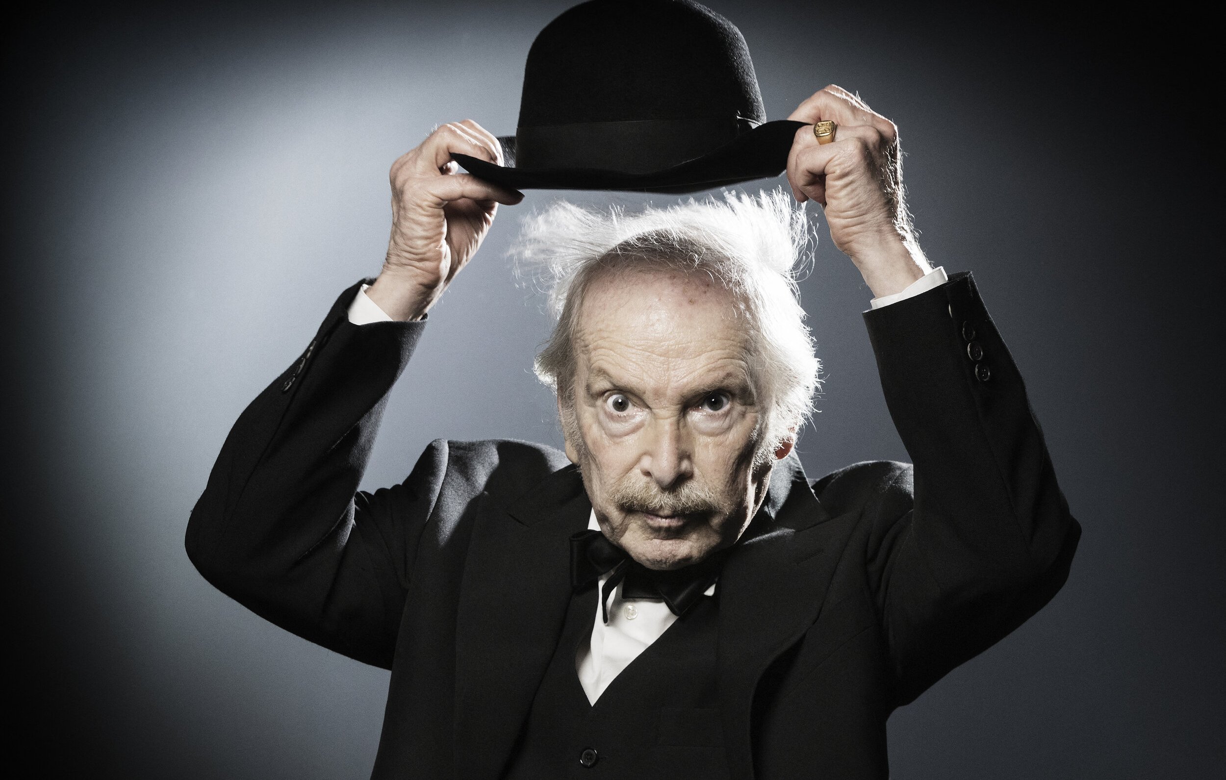 L'image montre un homme âgé portant un costume noir avec un nœud papillon. Il tient un chapeau melon avec une main, et son autre main semble légèrement levée. Ses cheveux sont ébouriffés, ce qui donne un aspect excentrique à son expression. L'arrière-plan est sobre, avec une lumière qui crée un effet dramatique, mettant en valeur son visage et son chapeau.