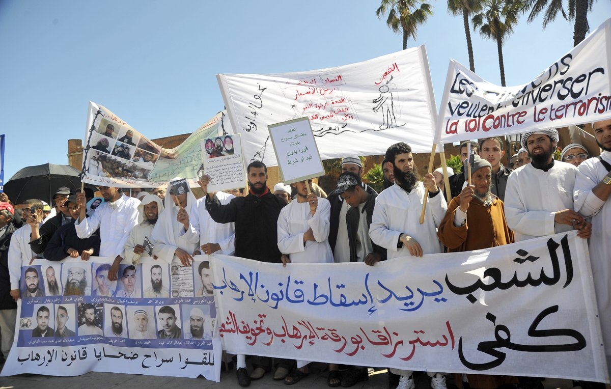 L'image montre une manifestation avec un groupe de personnes tenant des pancartes et des banderoles. Les manifestants semblent exprimer des revendications politiques ou sociales, avec des slogans écrits en arabe et en français. On peut voir des visages déterminés et des gestes de protestation. La scène est entourée d'arbres, suggérant un environnement urbain. L'atmosphère est chargée d'émotion, reflétant l'engagement des participants.