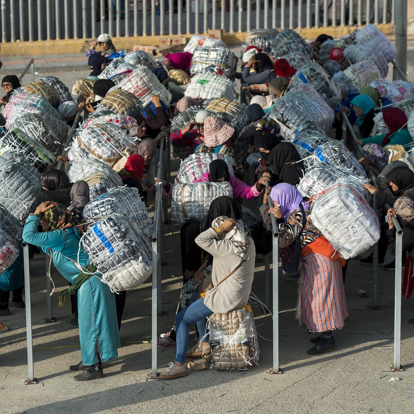 L'image montre un groupe de personnes, principalement des femmes, qui portent de grands fardeaux enveloppés dans des filets ou des tissus. Elles se trouvent dans un environnement urbain, probablement près d'un point de passage ou d'une frontière. La plupart des femmes sont vêtues de vêtements traditionnels et portent des foulards. Elles semblent attendre ou se regrouper, tandis qu'en arrière-plan, on aperçoit un soldat ou un garde. Cette scène évoque des thèmes de travail, de migration ou de transport de marchandises.