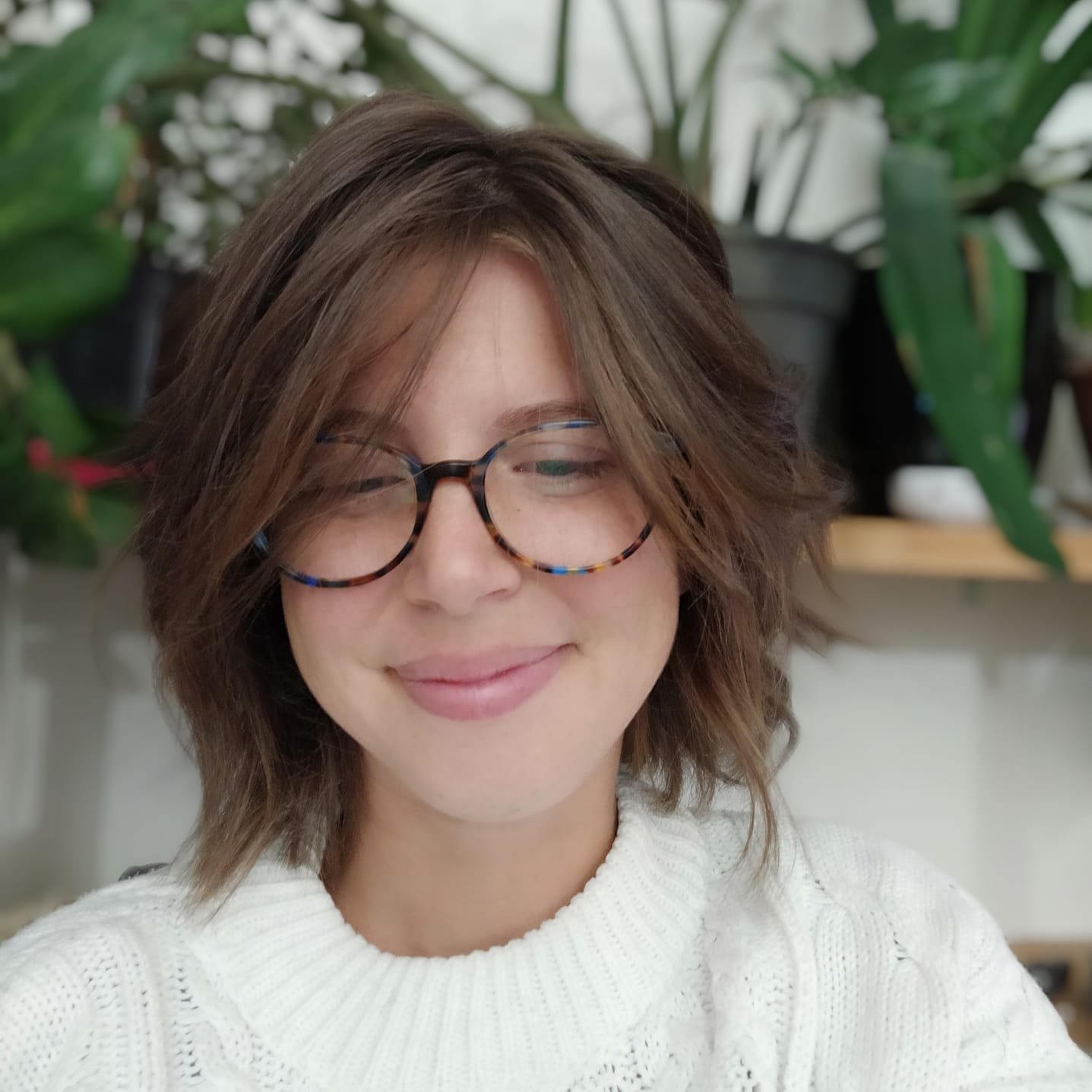 L'image montre une personne avec des cheveux bruns et légèrement ondulés, portant des lunettes à monture colorée. Elle sourit doucement et semble heureuse. Elle est vêtue d'un pull blanc en tricot. En arrière-plan, on peut apercevoir des plantes vertes, ce qui donne une ambiance naturelle et chaleureuse à la scène.