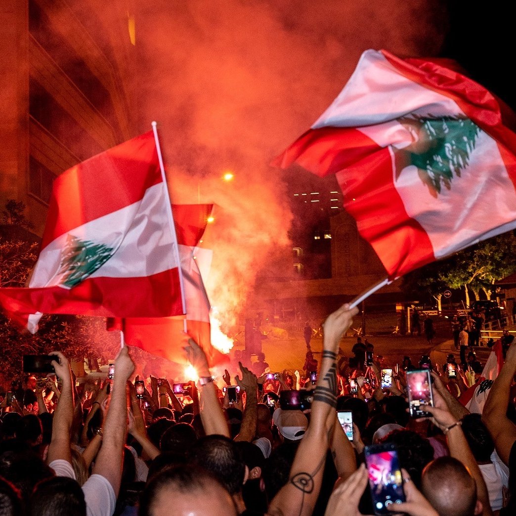 L'image montre une foule rassemblée, brandissant des drapeaux du Liban, dans une ambiance festive et animée. On peut voir des fumigènes rouges qui créent une atmosphère vibrante. De nombreuses personnes semblent capturer ce moment avec leurs téléphones. La scène se déroule probablement la nuit, avec des lumières urbaines en arrière-plan. L'engouement et la camaraderie parmi les participants sont palpables.