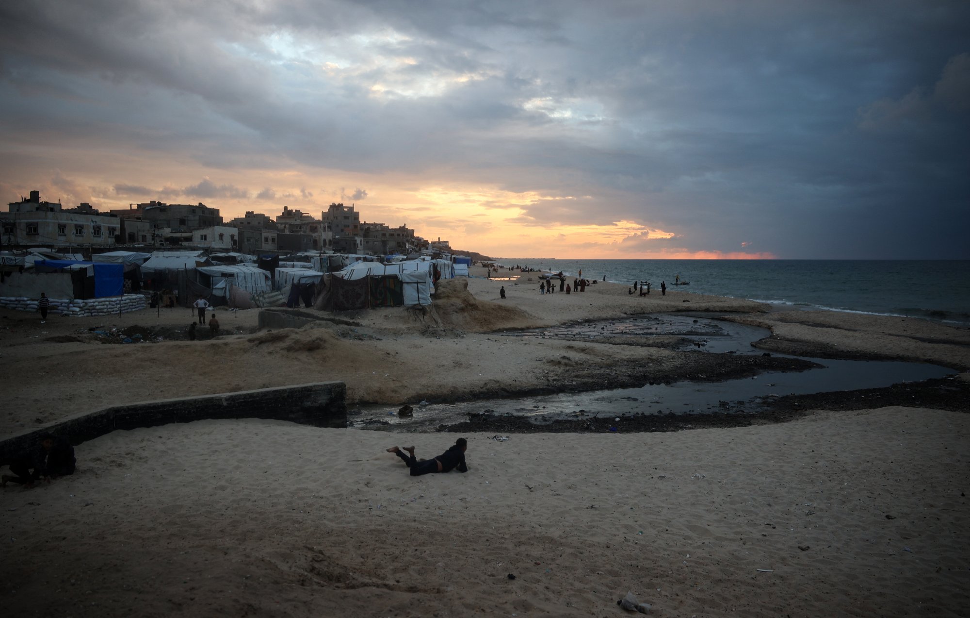 L'immagine mostra una spiaggia al tramonto, con il cielo che presenta nuvole scure e toni arancioni. Sulla spiaggia si trovano diverse tende e strutture, probabilmente utilizzate come rifugi temporanei. L'acqua del mare appare calma, e ci sono alcune persone in lontananza che camminano lungo la riva. In primo piano, si può vedere una parte di spiaggia con sabbia e un piccolo corso d'acqua che scorre. L'atmosfera sembrerebbe evocare un momento di transizione, sia dal punto di vista naturale che umano.