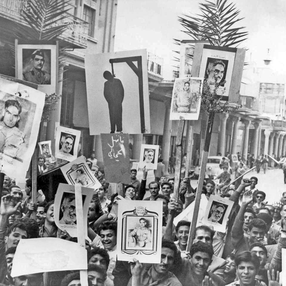 L'image montre une foule de personnes rassemblées dans une rue, tenant des pancartes et des portraits en protestation. On peut voir un dessin d'une potence au centre de l'affichage, suggérant un contexte de tension ou de révolte. Les visages des manifestants expriment diverses émotions, et l'environnement semble animé. Des palmiers sont visibles en arrière-plan, ajoutant une touche de verdure à la scène.
