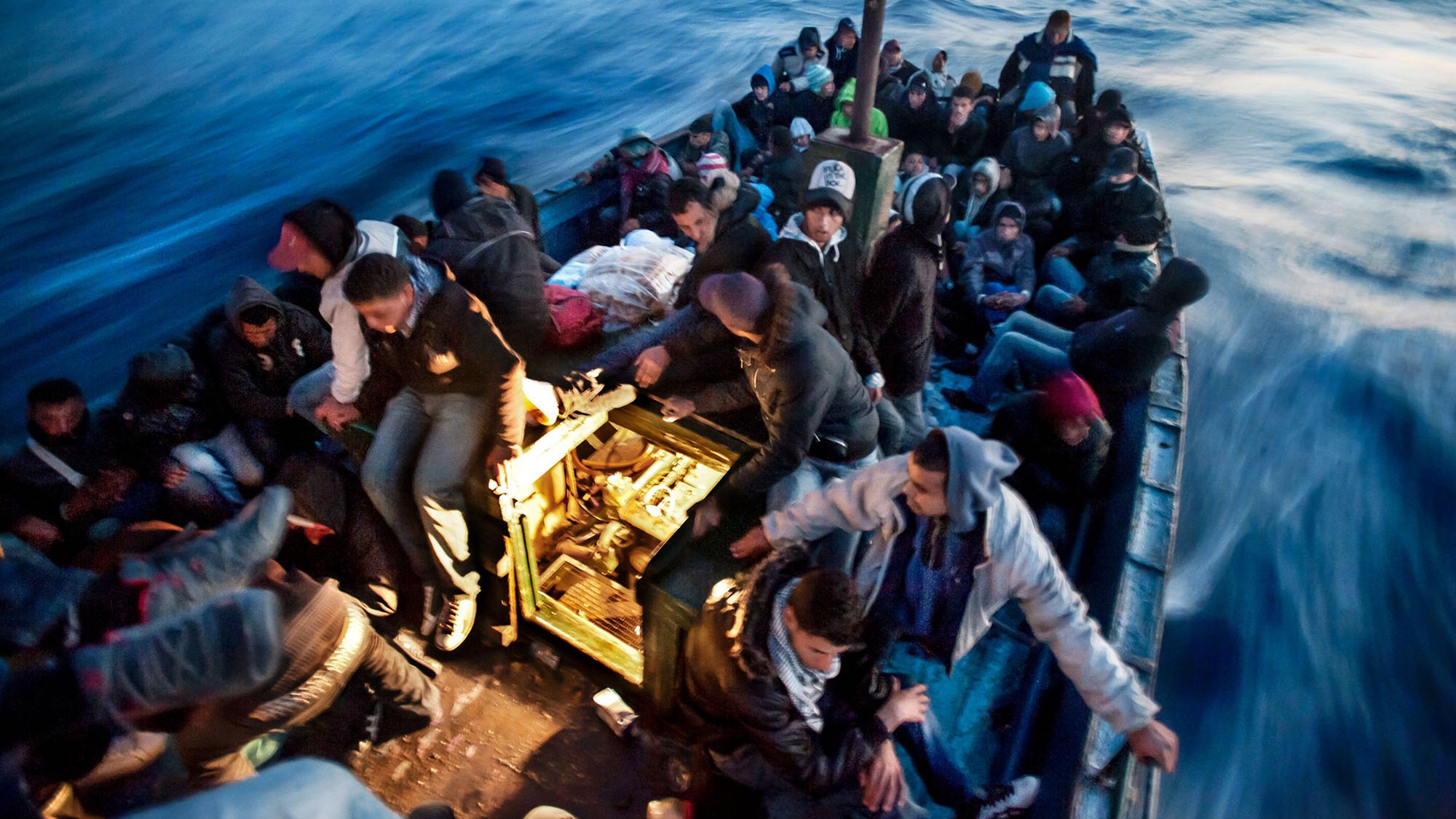 L'image montre une embarcation en mer, remplie de nombreuses personnes. Les passagers semblent être dans une situation difficile, tous étant assis ou debout, serrés les uns contre les autres. Ils portent des vêtements chauds, ce qui laisse supposer qu'ils font face à des conditions climatiques fraîches. L'eau est agitée et on peut voir des vagues, créant une atmosphère tendue et instable. La scène évoque des thèmes de migration et de recherche de refuge.