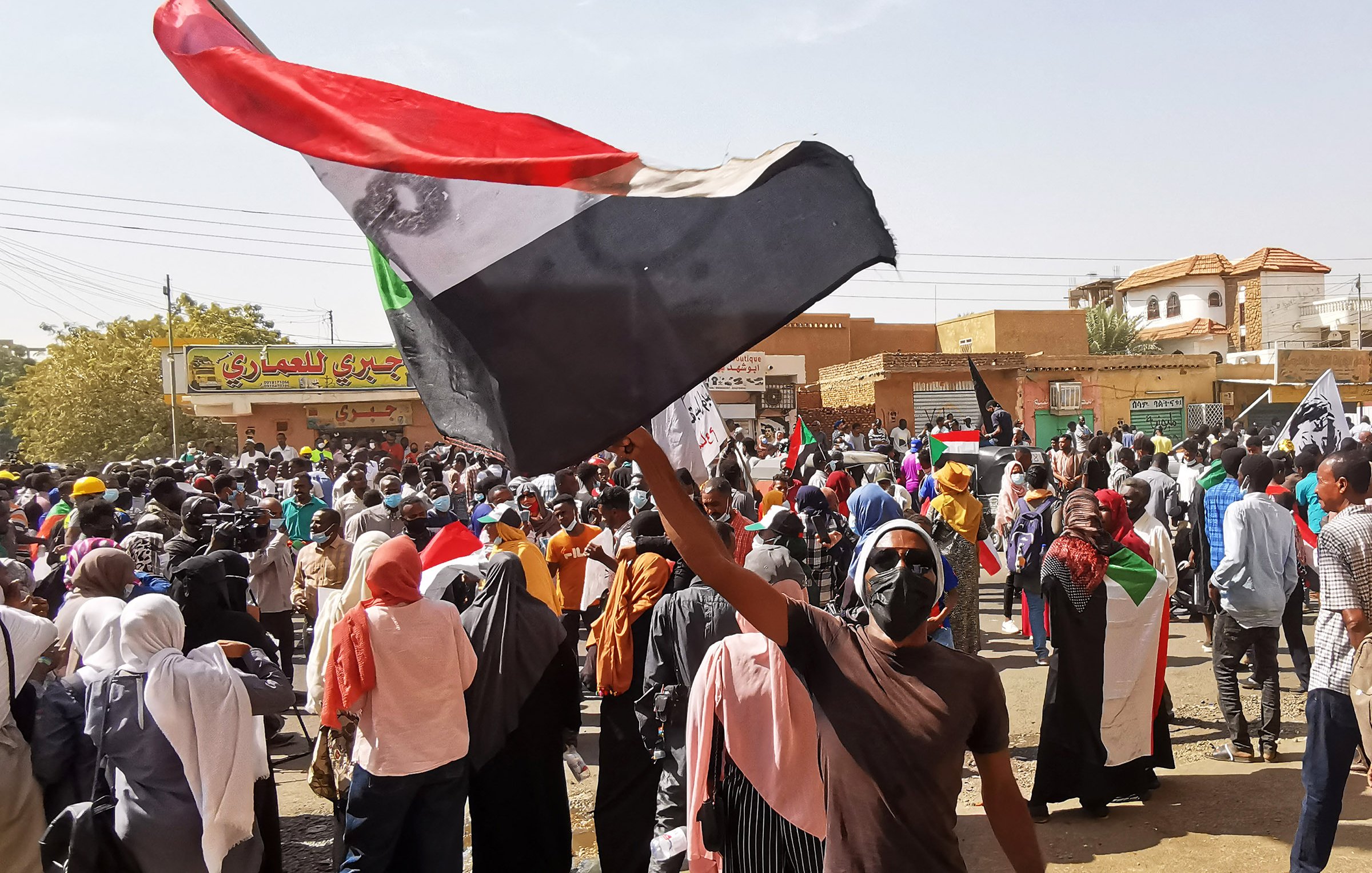 در این تصویر، یک گروه بزرگ از معترضان در خیابانی شلوغ جمع شده‌اند. فردی در پیش‌زمینه پرچم کشور سودان را به اهتزار درآورده و با شور و هیجان در میان جمعیت قرار دارد. مردم دیگری نیز در پس‌زمینه دیده می‌شوند که بعضی از آن‌ها پرچم‌ها و پلاکاردهایی در دست دارند. فضا به طور کلی بسیار پرنشاط و تحریک‌آمیز به نظر می‌رسد و نشان‌دهنده یک حرکت اعتراضی است. آفتاب روشن و آسمان آبی نیز بر جذابیت تصویر افزوده است.