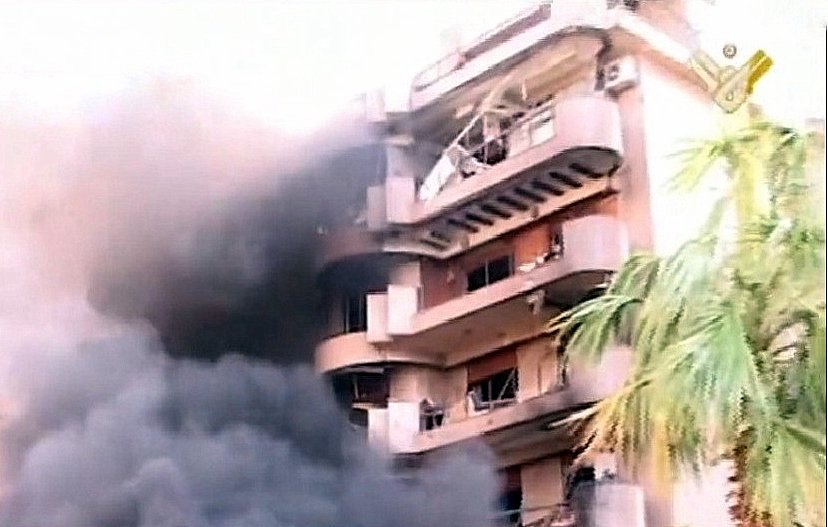L'image montre un bâtiment en proie à un incendie. De la fumée noire s'échappe des fenêtres, tandis que l'extérieur du bâtiment semble endommagé. Des éléments de végétation, comme un palmier, sont visibles au premier plan, contrastant avec la scène chaotique et la destruction. Le ton général de l'image est sombre, mettant en évidence l'urgence et la gravité de la situation.