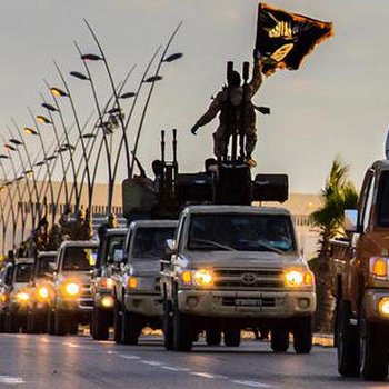 تظهر الصورة قافلة من الشاحنات تسير في شارع، حيث يوجد عدد من الأشخاص المسلحين على متنها. يُظهر جزء من القافلة لافتة سوداء تُشير إلى رمزية معينة. تبدو الأجواء في الصورة مائلة إلى المساء، مع وجود أعمدة إنارة على جوانب الطريق. توحي الصورة بحركة عسكرية أو تحركات لمجموعة معينة.