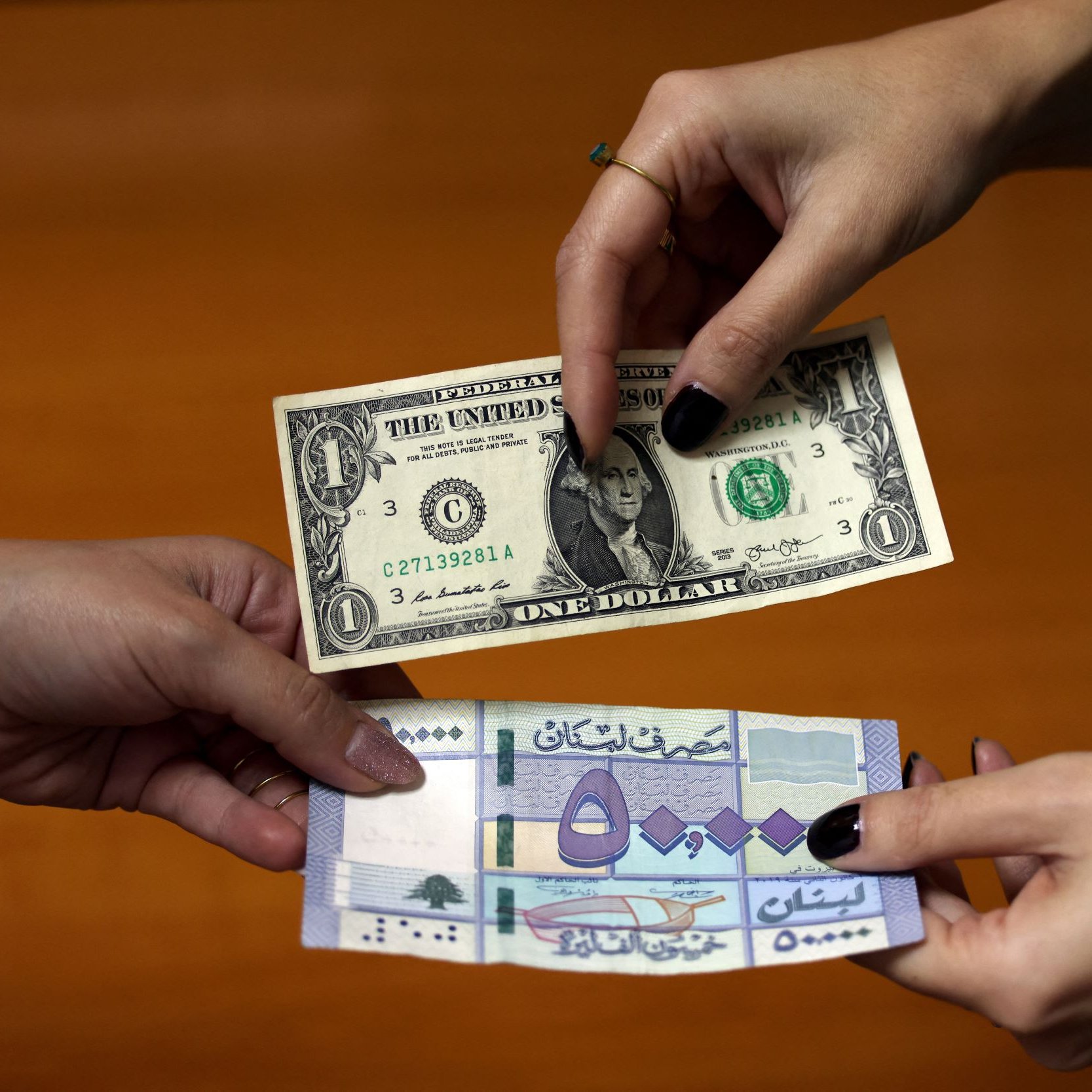 L'image montre deux mains tendant des billets de banque. À gauche, un billet d'un dollar américain est présenté, tandis qu'à droite, un billet d'une autre devise, probablement étrangère, est visible. Les deux billets sont tenus l'un près de l'autre, suggérant une transaction ou un échange monétaire. L'arrière-plan est flou, mettant l'accent sur les billets en question.