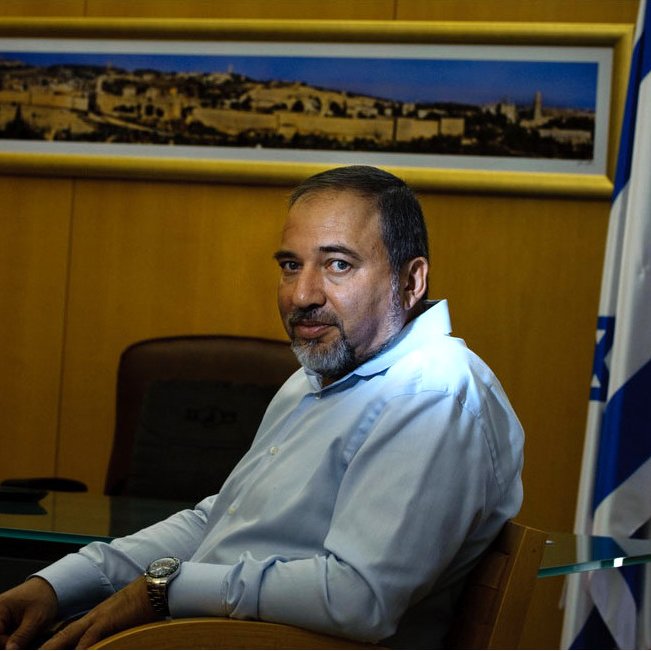 L'image montre un homme assis à un bureau. Il porte une chemise claire et a une expression sérieuse. En arrière-plan, on peut voir une grande photo d'une ville, probablement une vue de Jérusalem, et un drapeau, suggérant un contexte officiel ou politique. Le décor est sobre, avec une table en verre et une tasse sur le bureau.