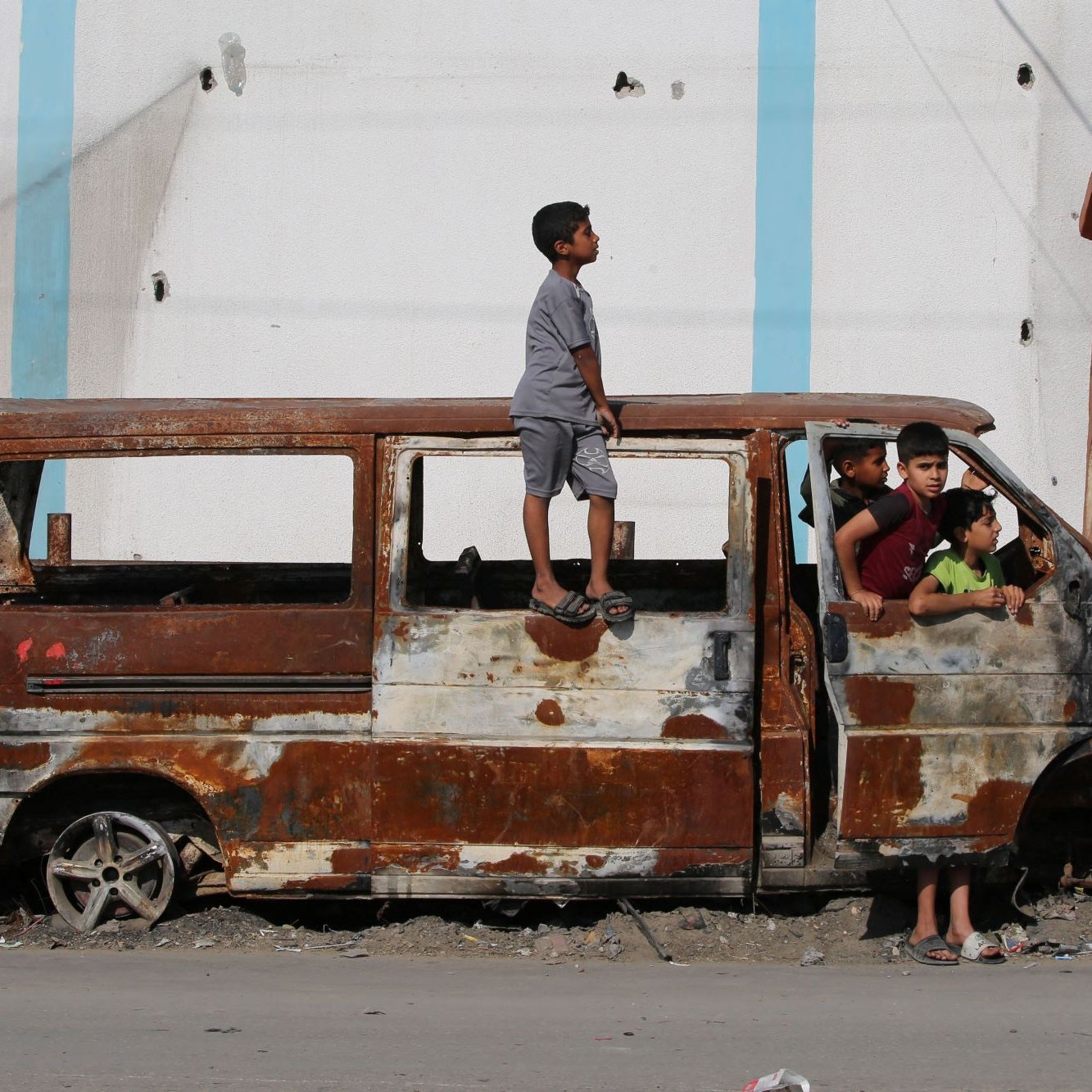 L'immagine mostra un gruppo di bambini che interagiscono attorno a un veicolo abbandonato e arrugginito. Uno dei bambini è in piedi sulla parte superiore di un furgone, mentre altri si trovano all'interno e intorno al veicolo. Lo sfondo evidenzia un edificio deteriorato, suggerendo un ambiente urbano trascurato. L'atmosfera sembra essere quella di un'area in difficoltà, ma i bambini sembrano divertirsi nonostante le condizioni.
