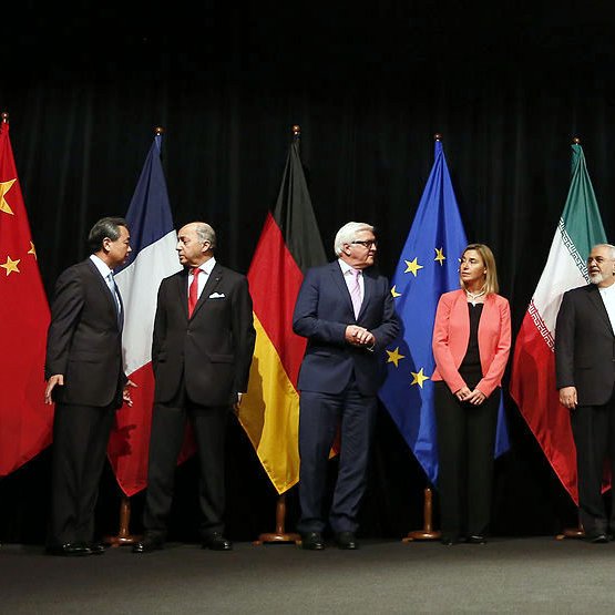 در این تصویر، گروهی از نمایندگان کشورهای مختلف در یک جلسه رسمی ایستاده‌اند. پرچم‌های چین، آلمان، اتحادیه اروپا، ایران، روسیه، بریتانیا و ایالات متحده در پس‌زمینه حضور دارند. هر یک از افراد در لباس‌های رسمی و با حالت‌های متفاوتی در صف ایستاده‌اند، که نشان‌دهنده اهمیت و جدیت جلسه است. به نظر می‌رسد آن‌ها در حال گفت‌وگو و تعامل با یکدیگر هستند.