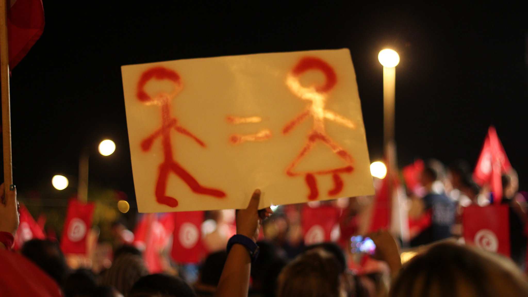 L'image montre une manifestation nocturne avec des gens rassemblés, portant des drapeaux rouges. Au premier plan, une personne tient une pancarte sur laquelle sont dessinées deux silhouettes : un homme et une femme, accompagnés du symbole "=" entre eux. Cela semble représenter un message d'égalité entre les sexes. L'ambiance est animée, et le public semble engagé sur le sujet.