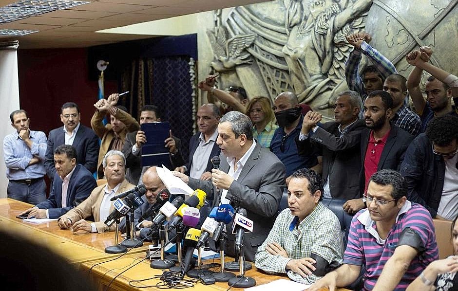 تظهر الصورة اجتماعًا تضامنياً أو مؤتمرًا صحفيًا حيث يجلس عدد من الأشخاص حول طاولة كبيرة، بينما يقف متحدث أمامهم يتحدث إلى الحضور. في الخلفية، يرفع آخرون أيديهم في إشارة إلى الاحتجاج أو التأييد. يظهر البعض في الصورة تفاعلهم مع المتحدث، مما يشير إلى أجواء حماسية. الخلفية تحتوي على منحوتات أو أنماط تزيينية، مما يضيف لمسة رسمية لمكان الاجتماع.