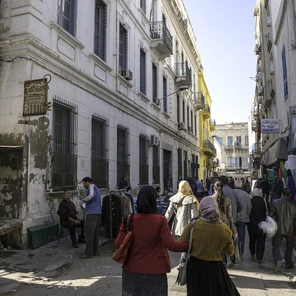 L'image montre une ruelle animée avec des bâtiments à l'architecture typique, dont certains sont peints en blanc et en jaune. On peut voir des gens marchant dans la rue, dont des femmes portant des habits traditionnels. À gauche, il y a un marché ou des stands avec des marchandises. L'ambiance semble vivante et typique d'un quartier urbain. La lumière du soleil éclaire la scène, ajoutant une chaleur à l'atmosphère.
