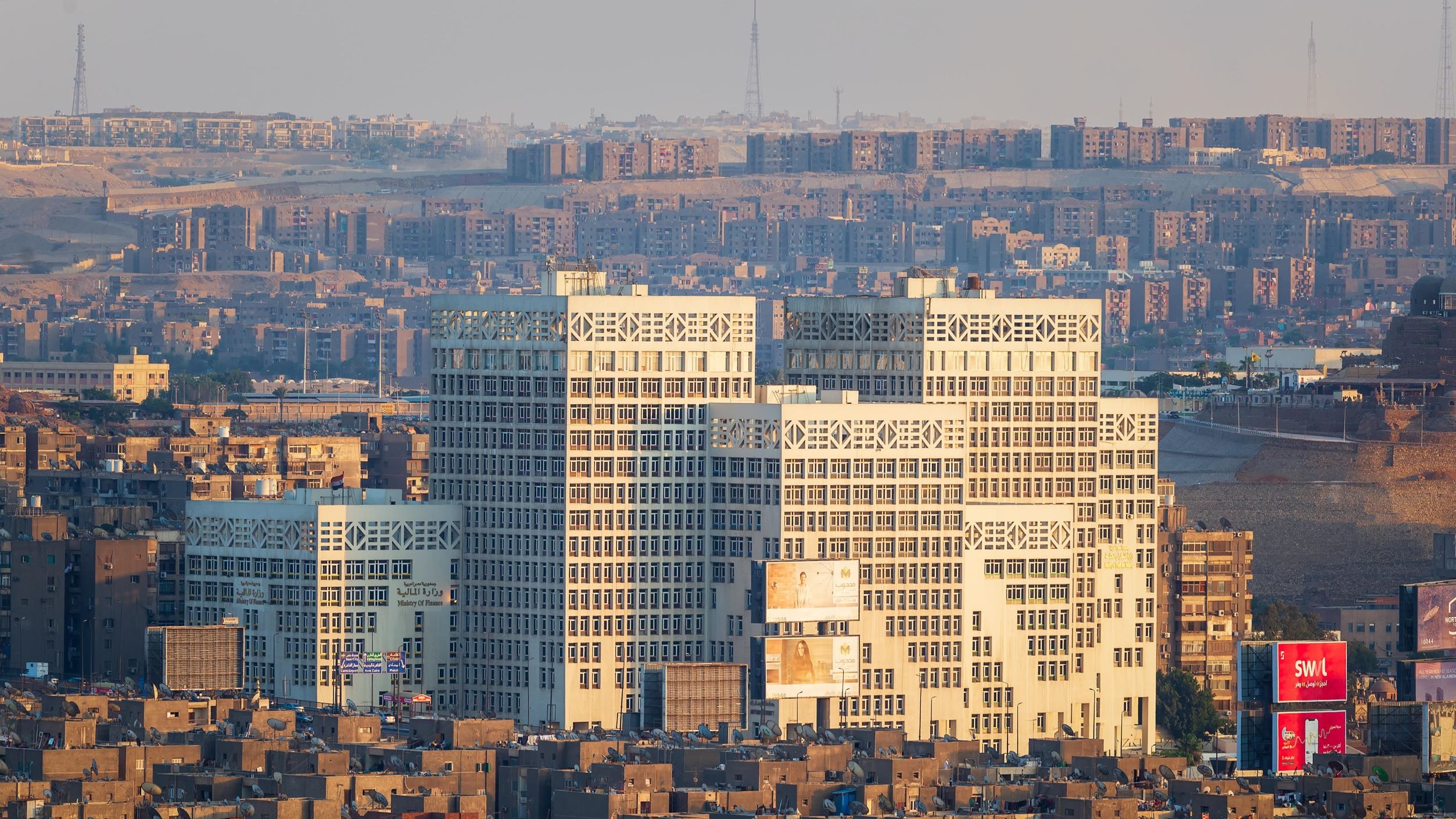 تظهر في الصورة مدينة تتميز بكثافة المباني والشقق السكنية. هناك مجموعة من المباني الحديثة ذات التصميم الفريد في المقدمة، حيث ينعكس عليها ضوء الشمس مما يبرز تفاصيلها. خلفها، تظهر منطقة سكنية كثيفة البناء، مع مشهد لجوانب مختلفة من المدينة. يبدو أن الجو هادئ، مع سماء مضيئة وقليل من الضباب.