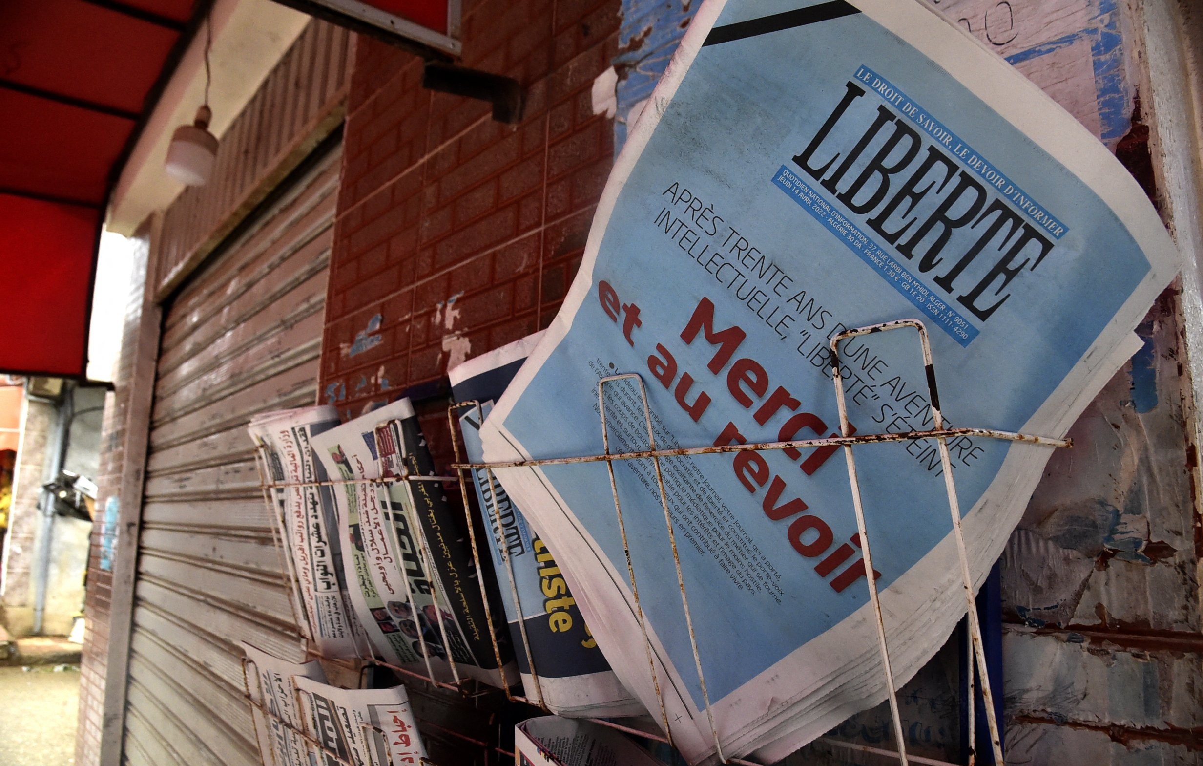 L'image montre un présentoir de journaux, où plusieurs titres sont visibles. Le journal en premier plan affiche le mot "LIBERTÉ" en lettres majuscules, accompagnés d'un sous-titre qui semble évoquer un sujet d'actualité. Les autres journaux à côté sont disposés de manière ordonnée, avec des titres variés, ce qui suggère un point de vente de presse dans un environnement urbain. Le mur en arrière-plan, ainsi que le toît rouge au-dessus, ajoutent à l'atmosphère locale de la scène.