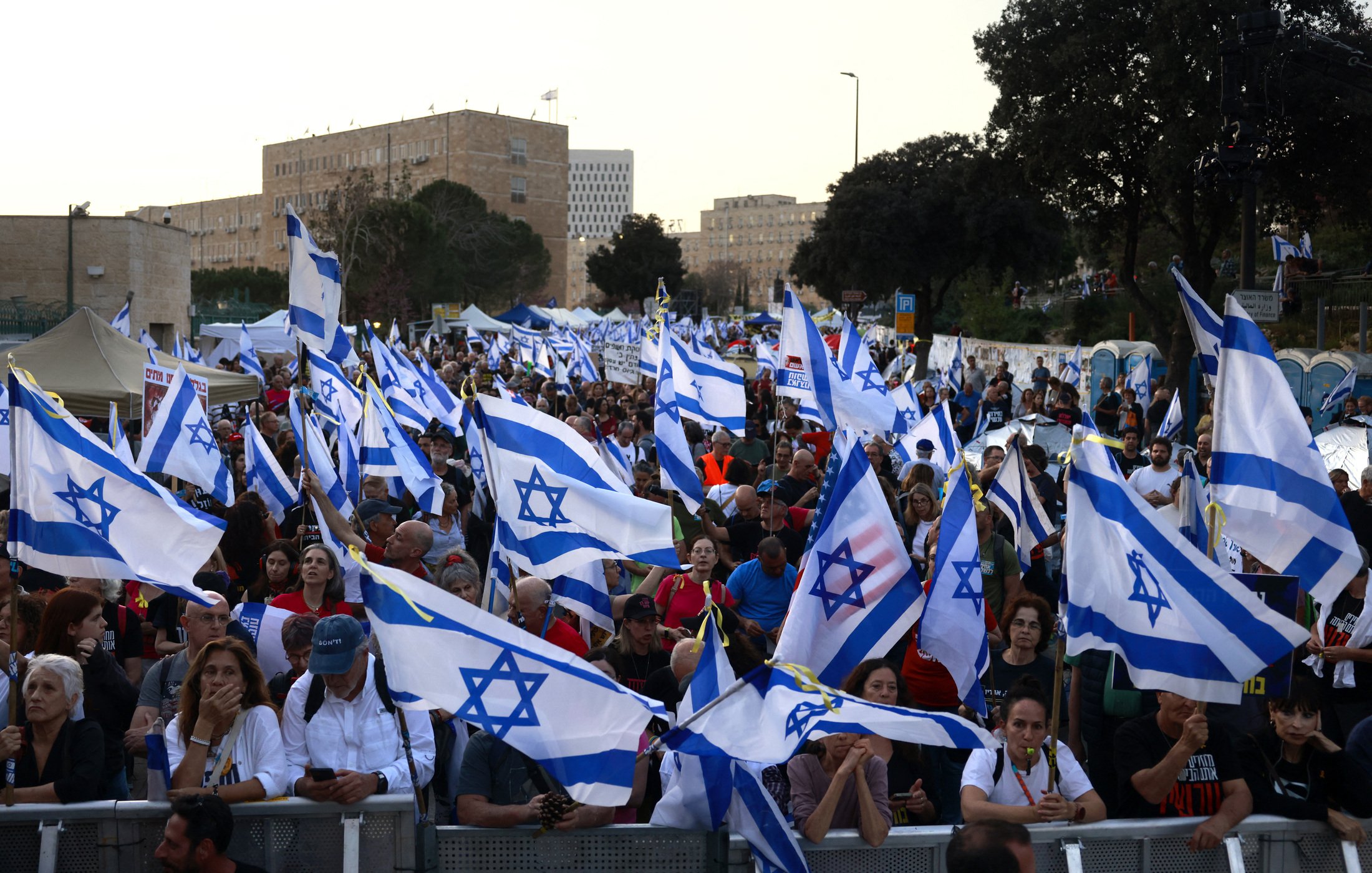 La imagen muestra una multitud de personas en una manifestación, ondeando banderas de Israel. La escena parece vibrante y llena de energía, con una gran cantidad de asistentes que levantan las banderas en un ambiente de protesta o celebración. En el fondo se pueden apreciar árboles y estructuras, lo que sugiere que el evento tiene lugar al aire libre, probablemente en una ciudad. La multitud parece diversa y está unida por una causa común.