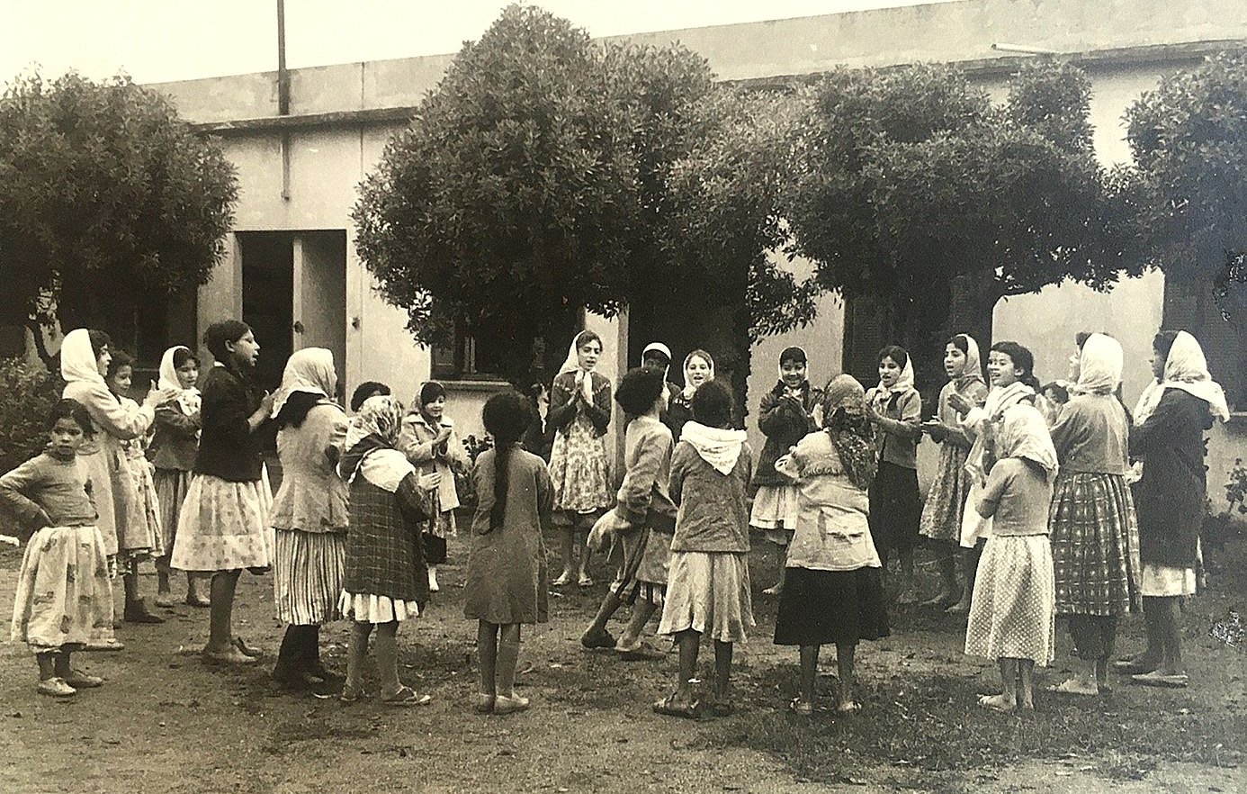 تظهر في الصورة مجموعة من الفتيات والأطفال يلعبون في ساحة مفتوحة. يبدو أنهن يرتدين ملابس تقليدية، وبعضهن يحمل غطاء رأس. الفتيات محاطة بأشجار، مما يضفي جوًا طبيعيًا على المكان. يبدون مشغولات باللعب والضحك، في مشهد يعكس روح الطفولة والنشاط. الخلفية بسيطة، مع وجود مبنى مكون من جدران بسيطة.